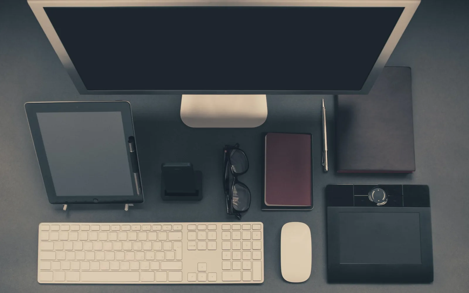 Top-down view of a developer desk with a monitor, tablet, keyboard, glasses and notebooks