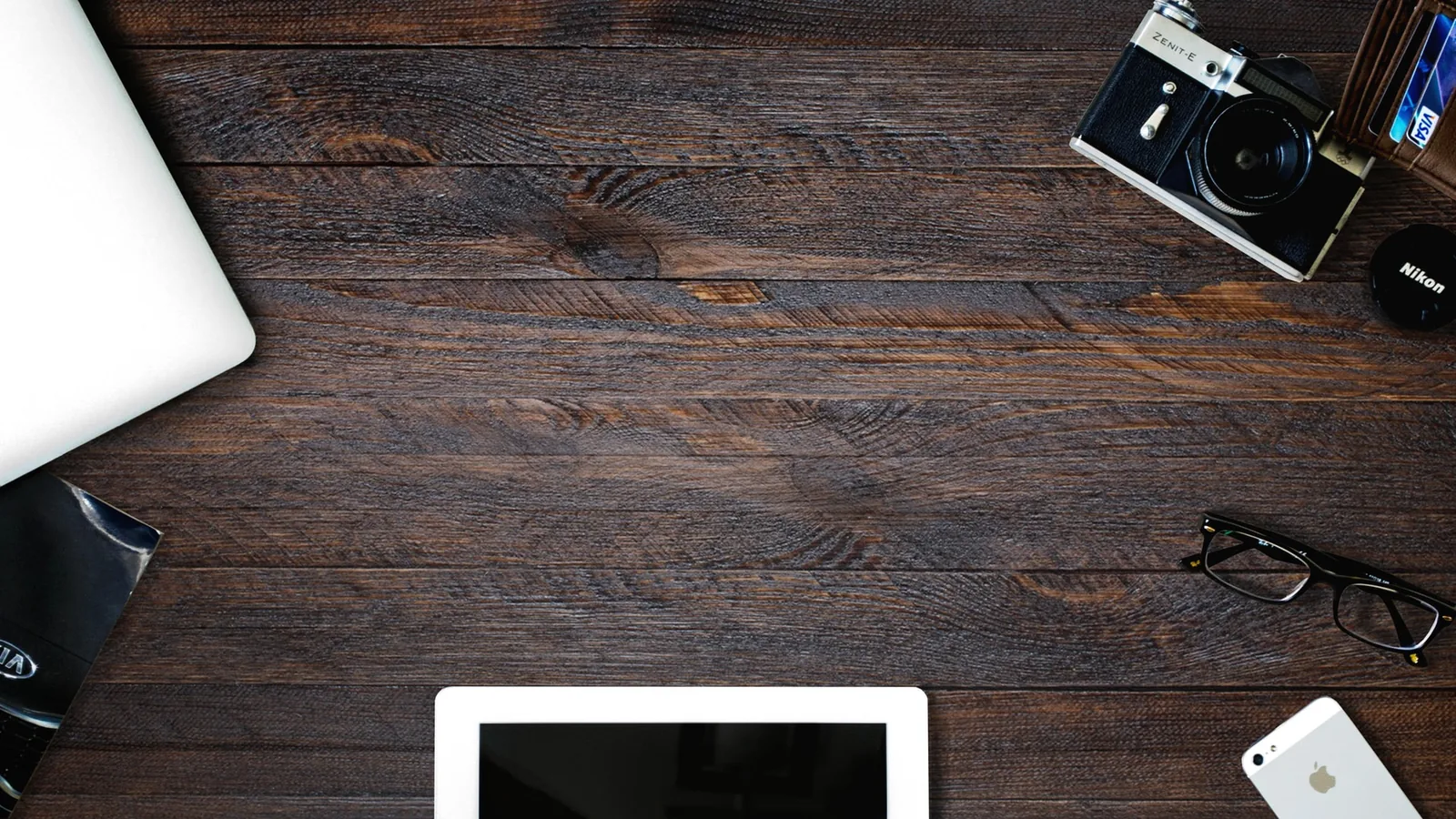 Dark wooden desk with a laptop, camera, glasses, tablet and phone around the edges